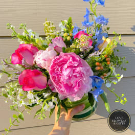 Bright bouquet of pink peonies, roses, and mixed blooms in a clear glass vase held in one hand