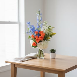Tall arrangement of orange roses, blue delphinium, and white hydrangeas in a glass vase on a wooden table.