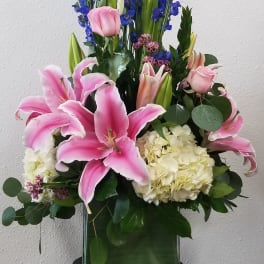 Tall floral arrangement with pink lilies, roses, blue delphinium, and white hydrangea in a glass vase