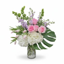 Tall glass vase arrangement with pink roses, tulips, white hydrangeas, lavender stock, and bold green leaves