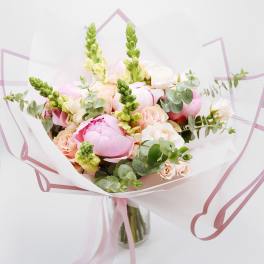 Hand-tied bouquet of pink peonies, peach roses, and snapdragons wrapped in white paper in a glass vase