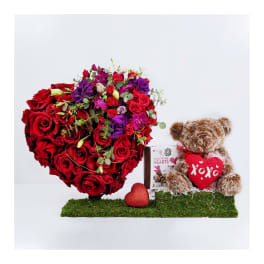 Heart-shaped red rose arrangement with a teddy bear and chocolates