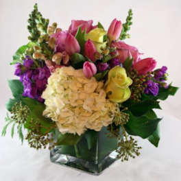Mixed bouquet with white hydrangea, pink tulips, yellow roses, and purple flowers in a clear glass cube vase