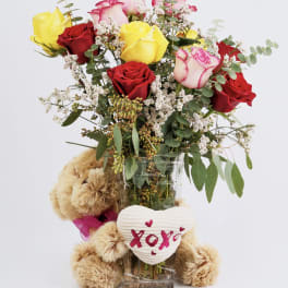 Bouquet of red and yellow roses in a clear vase with a teddy bear and heart decoration