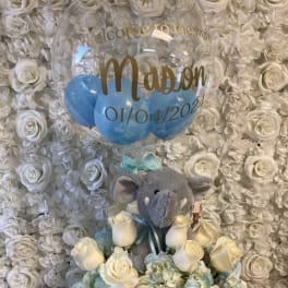 White rose arrangement with a gray plush elephant and blue balloons in a clear balloon