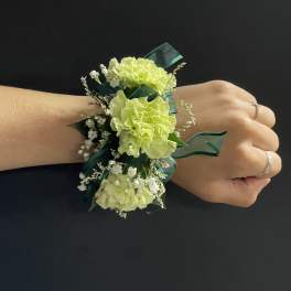 Light green carnation wrist corsage with white filler flowers and dark green ribbon on a hand.