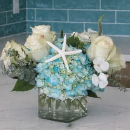 Blue hydrangea arrangement with white roses in a square glass vase and starfish accent