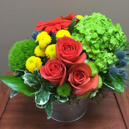 Compact arrangement of red roses, red gerbera, and mixed yellow and green blooms in a metal pot.