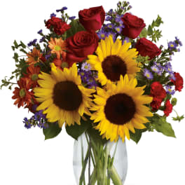 Vase arrangement of yellow sunflowers, red roses, orange daisies, and purple asters in a clear glass vase.