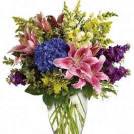 Mixed bouquet of pink lilies, blue hydrangea, and purple flowers in a clear glass vase