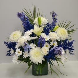 Tall arrangement of white lilies, roses, mums, and blue flowers in a green glass vase