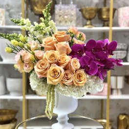 Peach roses and purple orchids in a white pedestal vase