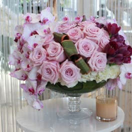 Pink roses and orchids arranged in a glass vase with a candle beside it.