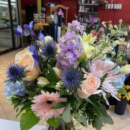Pastel mixed bouquet with roses, gerbera daisies, and blue accents arranged in a clear glass vase