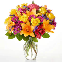 Mixed bouquet of yellow roses, orange lilies, and purple daisies in a glass vase