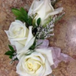 Corsage of three white roses with tiny white filler flowers and a pale pink ribbon bow.