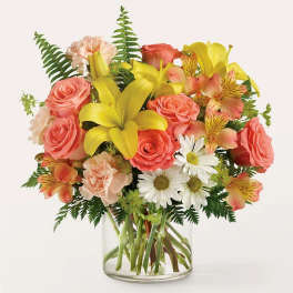 Mixed bouquet of yellow lilies, coral roses, and white daisies in a glass vase