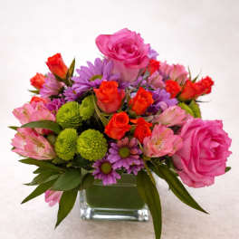 Mixed pink, purple, and orange flowers arranged in a square glass vase.