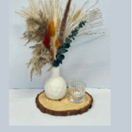 Dried floral arrangement in a white vase on a wood slice with a small glass candle holder