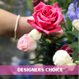 Florist hand holding a bright pink rose with small white and blush roses, labeled designers choice