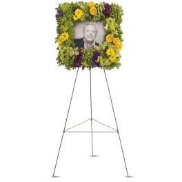 Floral memorial easel with a framed portrait surrounded by yellow, green, and purple flowers