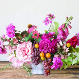 Bright pink floral arrangement with grapes and yellow berries in a white vase