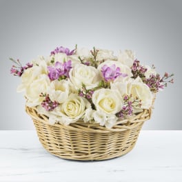 Low basket of white roses with light purple accent flowers in a woven wicker basket