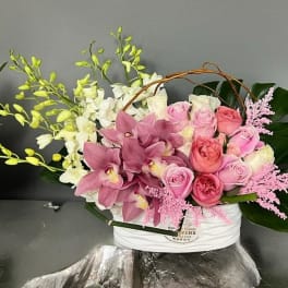 Pink roses and orchids arranged in a white basket with large green leaves