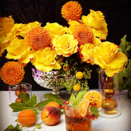 Yellow roses and orange dahlias arranged with fruit and glasses on a table.