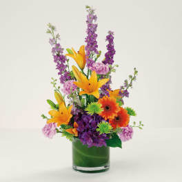 Tall mixed bouquet with orange lilies, purple blooms, and a green glass vase.