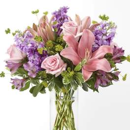 Pink lilies and roses in a clear glass vase with purple flowers