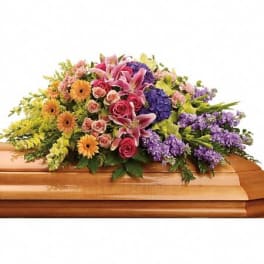 Colorful mixed flower casket spray with lilies, roses, and daisies on a wooden casket