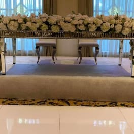 Long mirrored table wrapped in dense white and blush floral garlands on a stage.