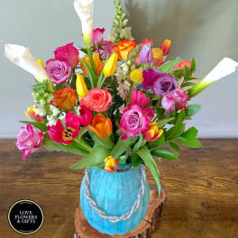 Colorful bouquet of roses, tulips, snapdragons, and calla lilies in a blue vase on a wood slab.
