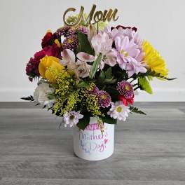 Mixed flower arrangement in a white Mother's Day container with a gold Mom topper