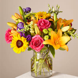 Mixed bouquet of roses, lilies, sunflowers, and purple blooms in a glass vase
