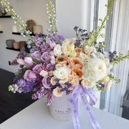 Lavender, peach, and white roses arranged in a round hatbox with a purple ribbon.