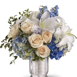 Arrangement of white lilies, cream roses, and blue hydrangeas in a clear glass vase