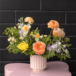 Low pastel arrangement of peach and yellow flowers in a ribbed vase on a pink table