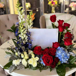 Floral arrangement of red roses, blue hydrangeas, and white blooms around a blank card