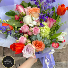 Hand-tied bouquet of mixed roses, tulips, and purple delphinium wrapped in lilac paper.
