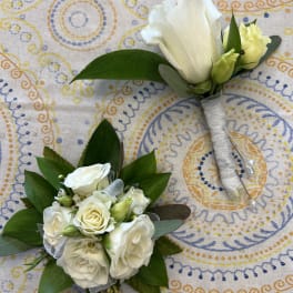 White rose boutonniere and matching corsage with ribbon accents on a patterned cloth.
