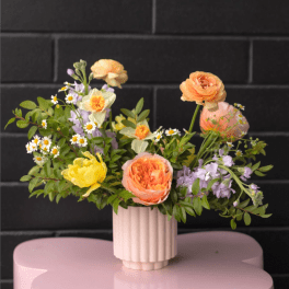 Low pastel arrangement of peach, yellow, and white flowers in a ribbed vase on a pink table