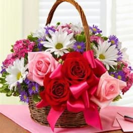 Wicker basket of red and pink roses with white daisies and purple flowers topped with a bright pink ribbon.