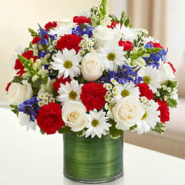Red, white, and blue mixed flower arrangement with roses, carnations, and daisies in a green-wrapped glass vase