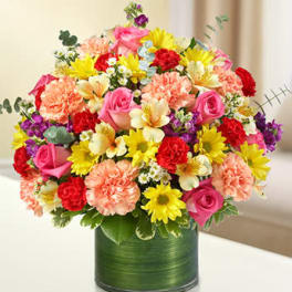 Colorful mixed bouquet of roses, carnations, daisies, and alstroemeria in a green-wrapped glass cylinder vase.
