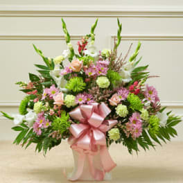 Fan-shaped arrangement of pink, green, and white flowers with a large pink bow in a white container.