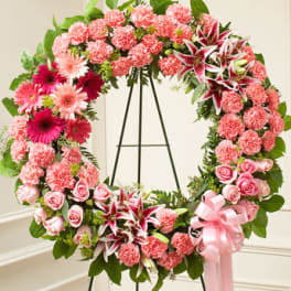 Large pink floral wreath with carnations, lilies, roses, and gerbera daisies on a metal easel with a pink bow