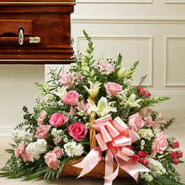 Pink and white funeral basket of roses, lilies, and carnations with bow beside a wooden casket