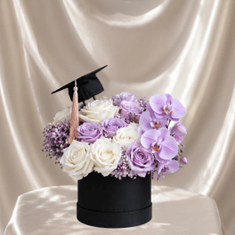 Lavender and white roses with purple orchids in a black box, topped with a graduation cap.